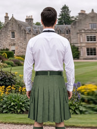 A man viewed from behind wearing a white dress shirt and an olive green pleated kilt, standing in a garden with a large stone mansion in the background.