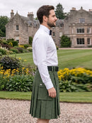 A man wearing a white dress shirt and a green pleated kilt stands in profile outdoors in front of a large stone mansion and manicured garden lawn.