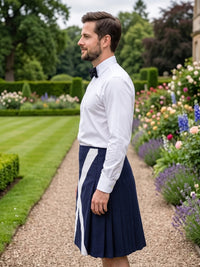A man standing in profile in a formal garden, wearing a white long-sleeve dress shirt, a black bow tie, and a navy blue pleated kilt with a white diagonal stripe.