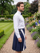 A man wearing a white long-sleeve dress shirt, black bow tie, and a navy blue kilt with a white diagonal stripe and silver buckle details, standing in a formal garden pathway.