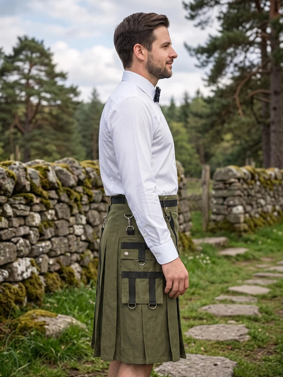 A man in profile wearing a white dress shirt, black bow tie, and an olive green kilt with black leather straps and buckles, standing outdoors on a stone path with a mossy stone wall and pine trees in the background.