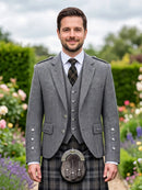 A man wearing a traditional Scottish formal outfit with a gray herringbone blazer, matching waistcoat, plaid kilt, and sporran, standing in a garden setting.
