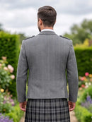 A man viewed from behind wearing a gray herringbone patterned jacket and a black and gray plaid kilt in a garden setting.