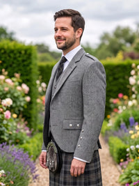 A man wearing a gray tweed blazer, white dress shirt, dark tie, and a black-and-gray plaid kilt with a sporran, standing in a lush garden pathway.