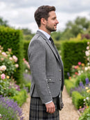 A man in profile wearing a gray herringbone blazer, a dark plaid kilt, a white shirt, and a tie, standing in a garden path with hedges and flowers in the background.