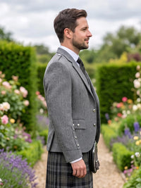 A man in profile wearing a gray herringbone blazer, a dark plaid kilt, a white shirt, and a tie, standing in a garden path with hedges and flowers in the background.