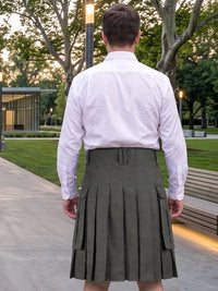 A man viewed from behind wearing a white long-sleeve dress shirt and an olive green pleated kilt in an outdoor park setting at dusk.