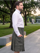 A man wearing a white dress shirt, black bow tie, and an olive green kilt with a front pocket, standing on a paved path in a park.