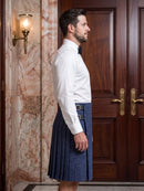 A man in profile wearing a white long-sleeve dress shirt and a dark blue pleated kilt with a matching waist belt, standing in front of a wooden door and marble wall.