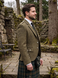 A man in profile wearing a brown tweed blazer, white dress shirt, dark tie, and a green tartan kilt with a black sporran, standing outdoors in front of stone ruins and trees.