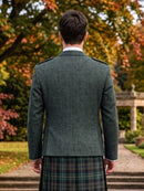 A man viewed from behind wearing a dark green tweed blazer and a green, blue, and black plaid kilt, standing outdoors on a stone path with autumn trees in the background.
