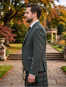 A man in profile wearing a dark green herringbone tweed blazer, a tartan kilt, and a sporran, standing outdoors on a stone path in a garden with autumn foliage.