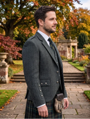 A man wearing a dark gray herringbone tweed kilt jacket and matching waistcoat, paired with a green and black patterned tie and a tartan kilt, standing in a stone garden path with autumn trees in the background.