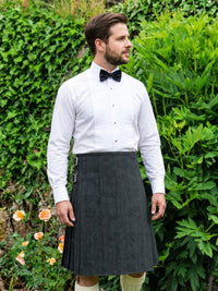 A man wearing a dark gray pleated kilt, a white dress shirt, and a black bow tie, standing outdoors in front of a green garden background.