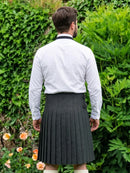 A man viewed from behind wearing a white long-sleeve dress shirt tucked into a dark gray pleated kilt, standing outdoors in front of a lush green garden background.