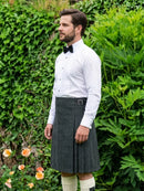 A man wearing a dark gray kilt, white dress shirt, black bow tie, and white knee socks, standing outdoors in front of a lush green ivy-covered wall.