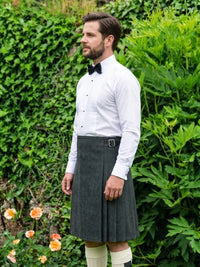 A man wearing a dark gray kilt, white dress shirt, black bow tie, and white knee socks, standing outdoors in front of a lush green ivy-covered wall.