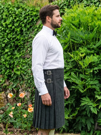 A man wearing a dark gray pleated kilt with double buckle straps, a white dress shirt, and a black bow tie, standing outdoors in front of a green garden wall.