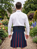 A man viewed from behind wearing a white dress shirt and a navy blue kilt with red stripes, standing in a garden path.