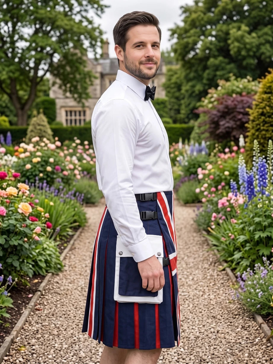 A man standing in a gravel garden path wearing a navy blue kilt with red and white stripes, a white dress shirt, and a black bow tie. He is holding a white tablet case.