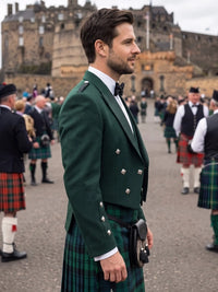 A man in a traditional Scottish formal outfit, featuring a dark green double-breasted jacket, a green tartan kilt, a white shirt, and a black bow tie, standing outdoors in front of Edinburgh Castle with other men in kilts in the background.