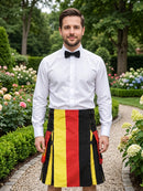 A man standing in a garden wearing a white dress shirt, black bow tie, and a pleated kilt with vertical black, red, and yellow stripes resembling the German flag.