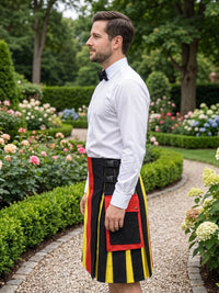 A man in a white dress shirt and black bow tie stands in a garden wearing a traditional Scottish kilt with black, red, and yellow stripes and a black waist belt.