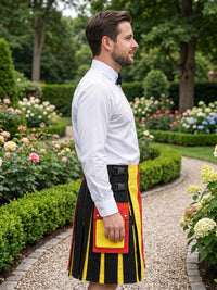A man in a white dress shirt and black bow tie stands in profile in a garden, wearing a black kilt with bold vertical yellow and red stripes and matching striped pockets.