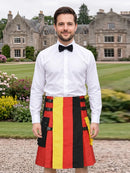 A man wearing a white dress shirt, black bow tie, and a red, yellow, and black striped kilt stands in front of a large stone mansion on a green lawn.