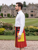A man wearing a white dress shirt, black bow tie, and a red kilt with black and yellow accents, standing in a garden with a large stone mansion in the background.