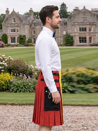 A man in a white dress shirt and black bow tie stands in profile outdoors in front of a large stone mansion and manicured lawn, wearing a red pleated kilt with black and yellow vertical panels and a black sporran pouch.