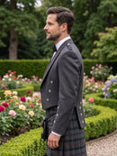 A man in profile wearing a dark gray kilt jacket and a tartan kilt, standing in a lush garden with flowers and green hedges in the background.