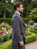 A man in profile wearing a dark gray formal jacket and a green plaid kilt, standing in a landscaped garden with trimmed hedges and colorful flowers.