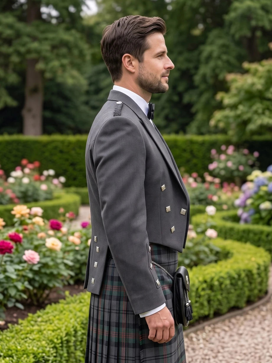 A man in profile wearing a dark gray formal jacket and a green plaid kilt, standing in a landscaped garden with trimmed hedges and colorful flowers.