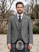 A man wearing a traditional Scottish formal outfit with a gray herringbone blazer, matching waistcoat, white shirt, patterned tie, and a gray plaid kilt with a sporran, standing outdoors in front of a stone wall and bare trees.