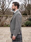 A man in profile wearing a gray herringbone blazer and a black-and-gray plaid kilt, standing outdoors on a stone patio with bare trees and a stone wall in the background.