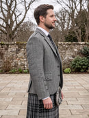 A man in profile wearing a gray herringbone tweed blazer, a black and gray plaid kilt, a white dress shirt, and a dark tie, standing outdoors on a stone paved area with a stone wall and bare trees in the background.