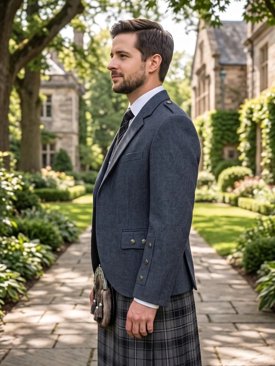 A man in profile wearing a dark navy blazer, white dress shirt, black tie, and a gray plaid kilt with a sporran, standing on a stone path in a garden courtyard with stone buildings in the background.