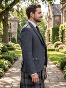 A man in profile wearing a dark gray blazer, white dress shirt, black tie, and a gray plaid kilt with a leather sporran, standing on a stone path in a garden courtyard with stone buildings in the background.