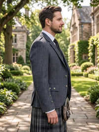 A man in profile wearing a dark gray blazer, white dress shirt, black tie, and a gray plaid kilt with a leather sporran, standing on a stone path in a garden courtyard with stone buildings in the background.