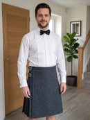 A man wearing a white long-sleeve pleated shirt, black bow tie, and a dark gray kilt with silver buckle details, standing in a modern home hallway.
