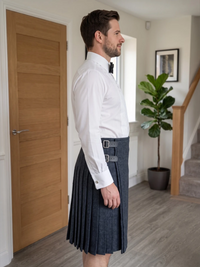 A man wearing a dark gray pleated kilt with a matching belt and silver buckle, paired with a white dress shirt and black bow tie, standing in profile in a modern home hallway.