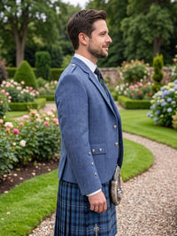 A man in profile wearing a blue kilted jacket and a blue tartan kilt, standing on a gravel path in a lush green garden.