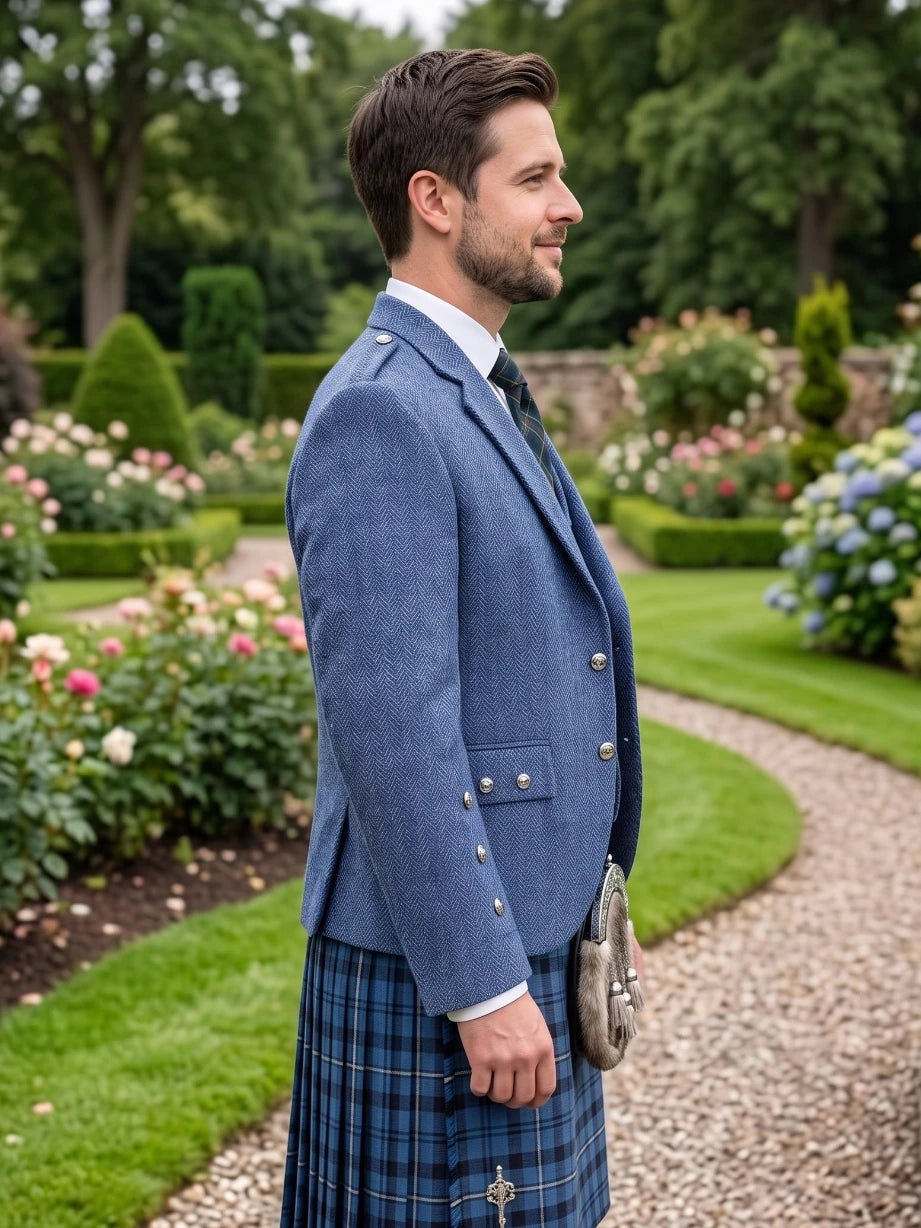 A man in profile wearing a blue kilted jacket and a blue tartan kilt, standing on a gravel path in a lush green garden.