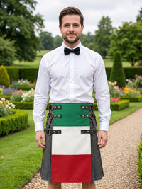 A man wearing a white dress shirt, black bow tie, and a traditional Scottish kilt with the green, white, and red vertical stripes of the Italian flag, standing in a manicured garden.