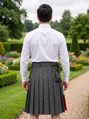 A man viewed from behind wearing a white dress shirt and a dark gray pleated kilt with black and gold buckle details, standing on a garden path.