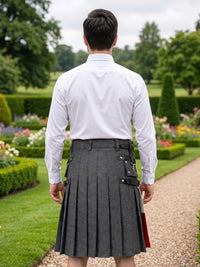 A man viewed from behind wearing a white dress shirt and a dark gray pleated kilt with black and gold buckle details, standing on a garden path.