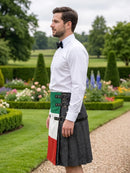 A man standing in a formal garden wearing a white dress shirt, black bow tie, and a kilt with an Irish flag pattern and a black leather strap system.