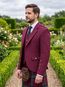 A man wearing a burgundy tweed kilt blazer, white dress shirt, dark tie, and a red tartan kilt with a brown sporran, standing in a garden path.