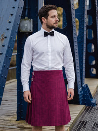 A man wearing a white dress shirt, black bow tie, and a magenta pleated kilt with silver buckle details, standing on a wooden walkway in front of a blue steel bridge.